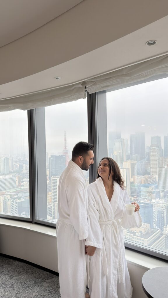 Couple in bathrobes beside tall windows with sheer curtains and a city view at Park Hotel Tokyo
