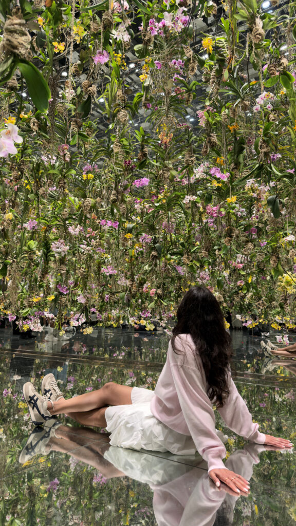 Woman reclining on the mirrored floor beneath thousands of hanging orchids in the Floating Flower Garden at teamLab Planets, Tokyo