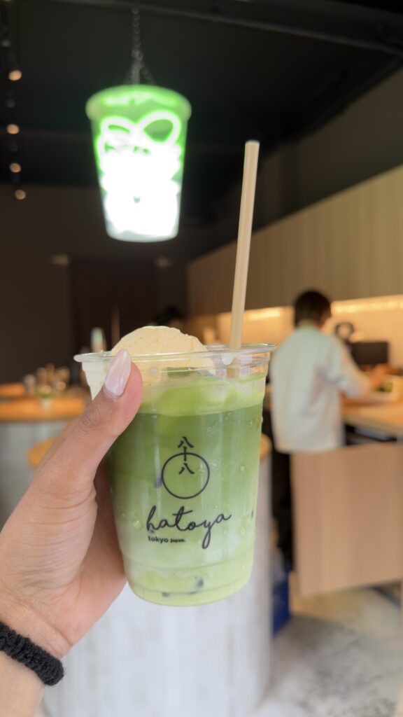 Hand holding a matcha latte in a Hatoya cafe cup beneath a neon matcha-cup light in Shinjuku, Tokyo